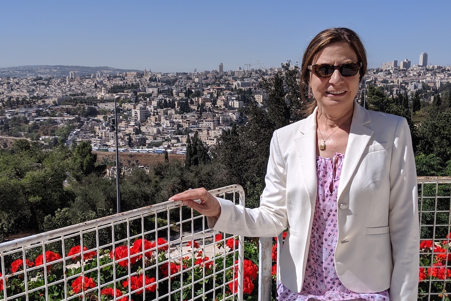 woman with sunglasses wearing a white jacket and pink top standing in front of a city