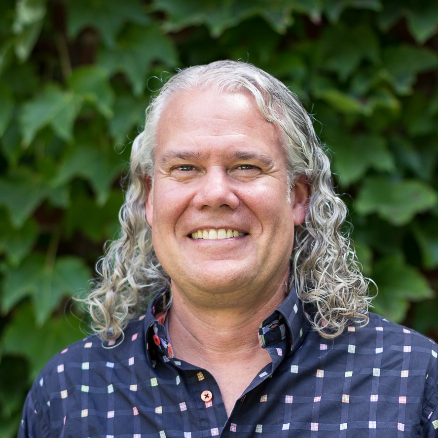 headshot of a man with long white hair wearing a blue button down shirt