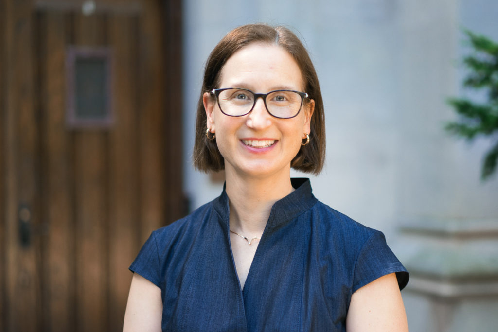 A picture of a woman with short brown hair and glasses wearing a blue short-sleeve shirt. 