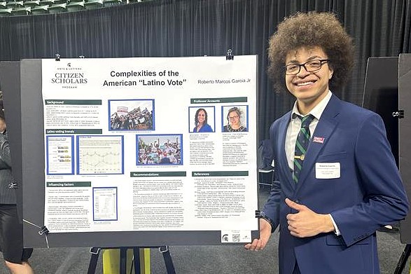 A student smiling and wearing glasses and a blue suit stands beside a large academic poster titled “Complexities of the American ‘Latino Vote.’”
