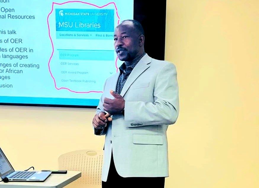 A man giving a presentation. He is wearing a light gray blazer and standing in front of a large screen and yellow wall. A laptop and desk are visible in the foreground.