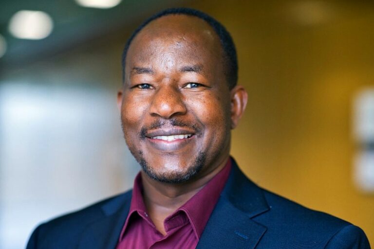 A man smiling in a close-up portrait. He is wearing a dark suit jacket and a maroon shirt.