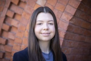 A woman with long, straight brown hair smiles at the camera in front of a brick wall. She wears a navy blazer and light blue shirt.