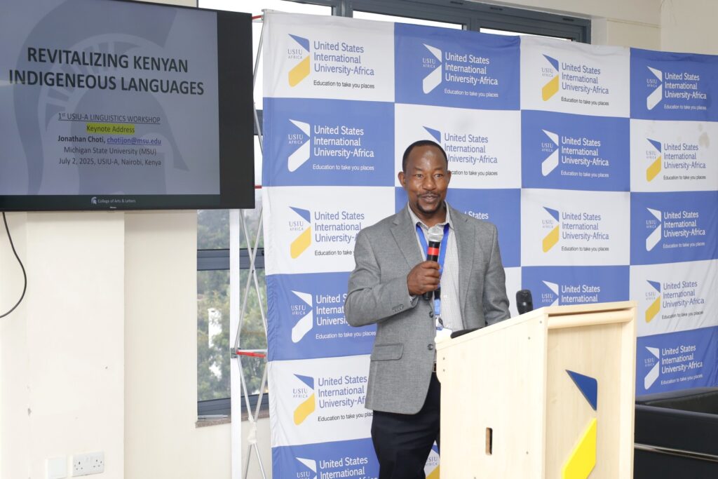 Black man standing at a podium and holding a microphone. He is wearing a gray suit. He is standing in front of a screen that says United States International University–Africa. He also is standing near a TV screen that say "Revitalizing Kenyan Indigeneous Languages.