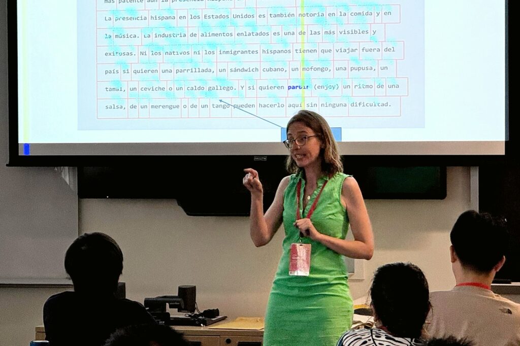 A woman stands in front of a classroom, gesturing while speaking to an audience. She wears a green dress and a red lanyard, and behind her is a projection of Spanish text on a screen. Several people are seated facing her, listening.
