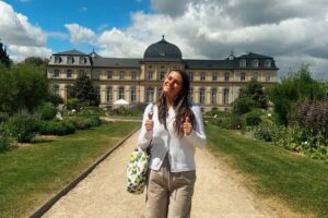 Emma Steury stands smiling with both thumbs up on a gravel path leading to a grand yellow and gray Baroque-style building in Germany. She is wearing light pants and a white top, with a patterned tote bag on her shoulder. The path is flanked by manicured gardens, and the sky is partly cloudy.