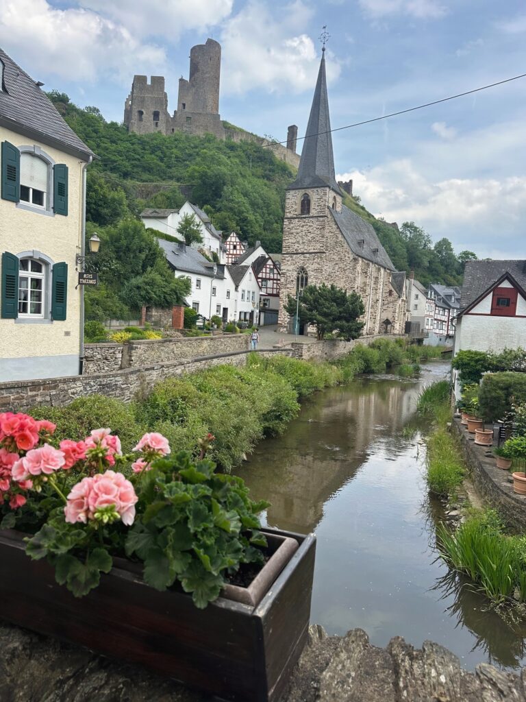 A scenic view of a small village in Germany, with a narrow stream running through the center. On the left, a planter box of pink flowers sits atop a stone bridge. Traditional half-timbered houses and a tall stone church with a pointed steeple line the banks, while medieval castle ruins rise on a hill in the background.