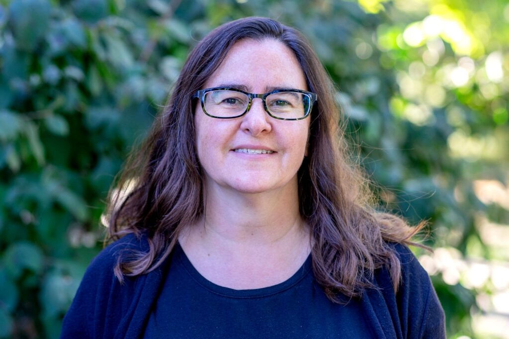 Headand shoulders photo of a woman with long brown and black framed glasses, smiling and wearing a dark blue shirt. She is standing in front of an area with green foliage. 
