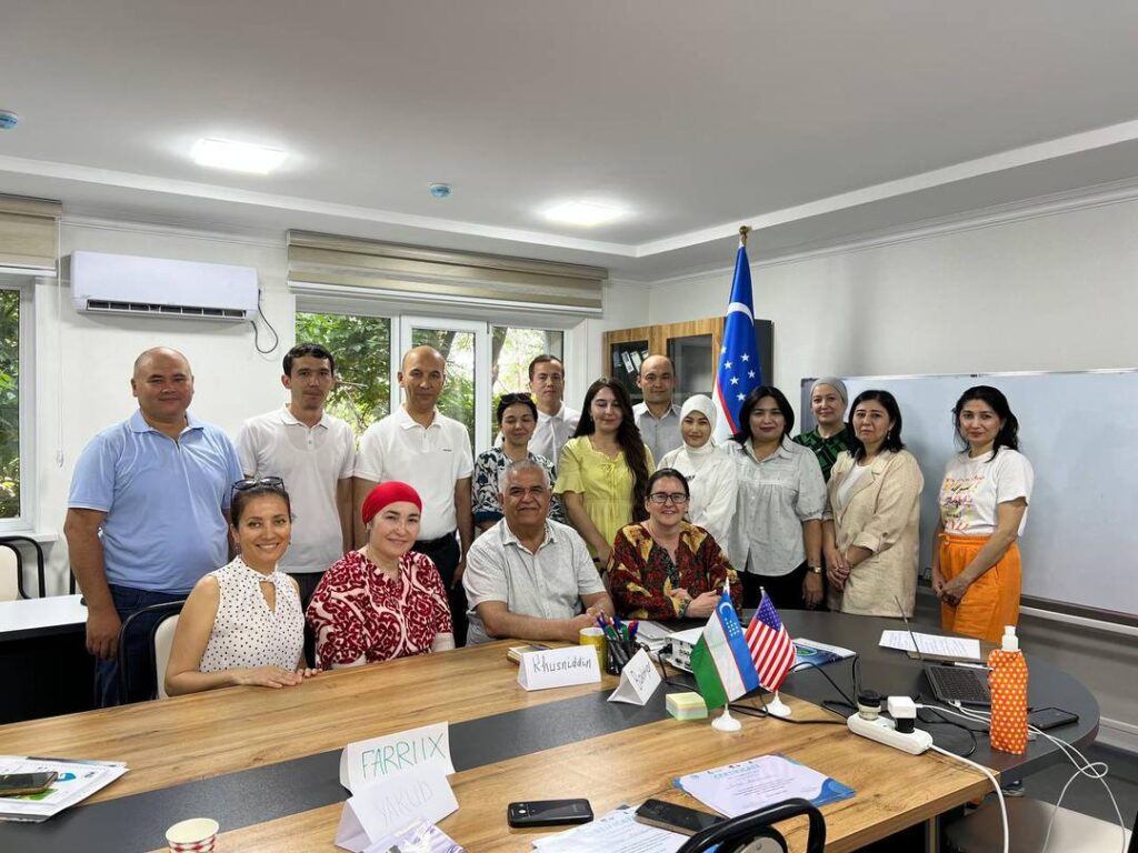A group of sixteen people, including men and women, pose for a photo in a conference room. Name cards, notebooks, and small flags of the U.S. and Uzbekistan are on the table in front of them.