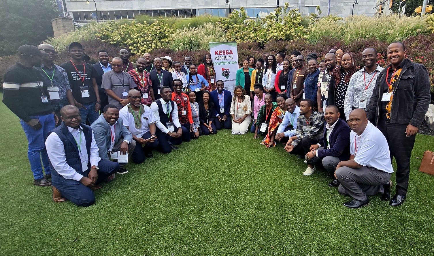 Group photo of 48 people, standing outside on a lawn with bushes and ornamental grass behind them. At the center of their gathering is a sign that says: "KESSA Annual Conference: Kenya Scholars & Studies Association." 