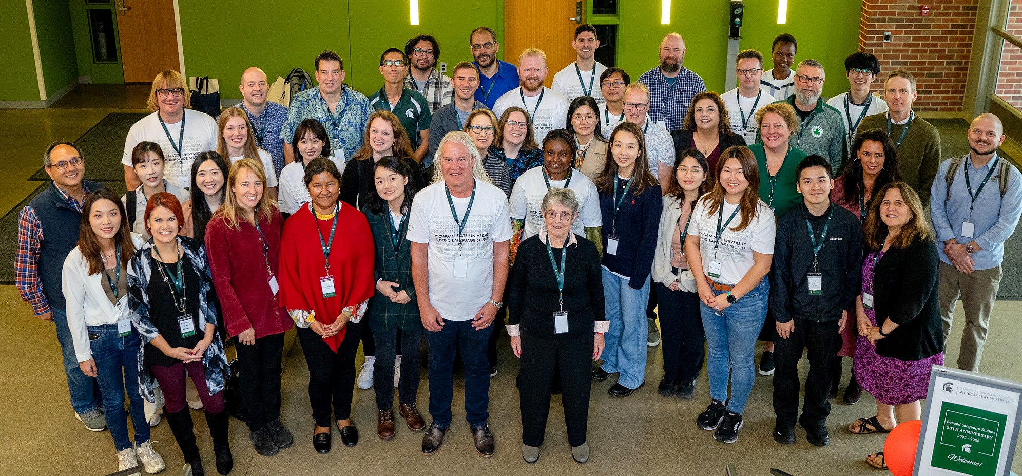 A large, diverse group of faculty, alumni, students, and guests pose together in a bright atrium with green walls during Michigan State University’s Second Language Studies Ph.D. program 20th anniversary celebration. Many wear MSU name badges and SLS-branded shirts.
