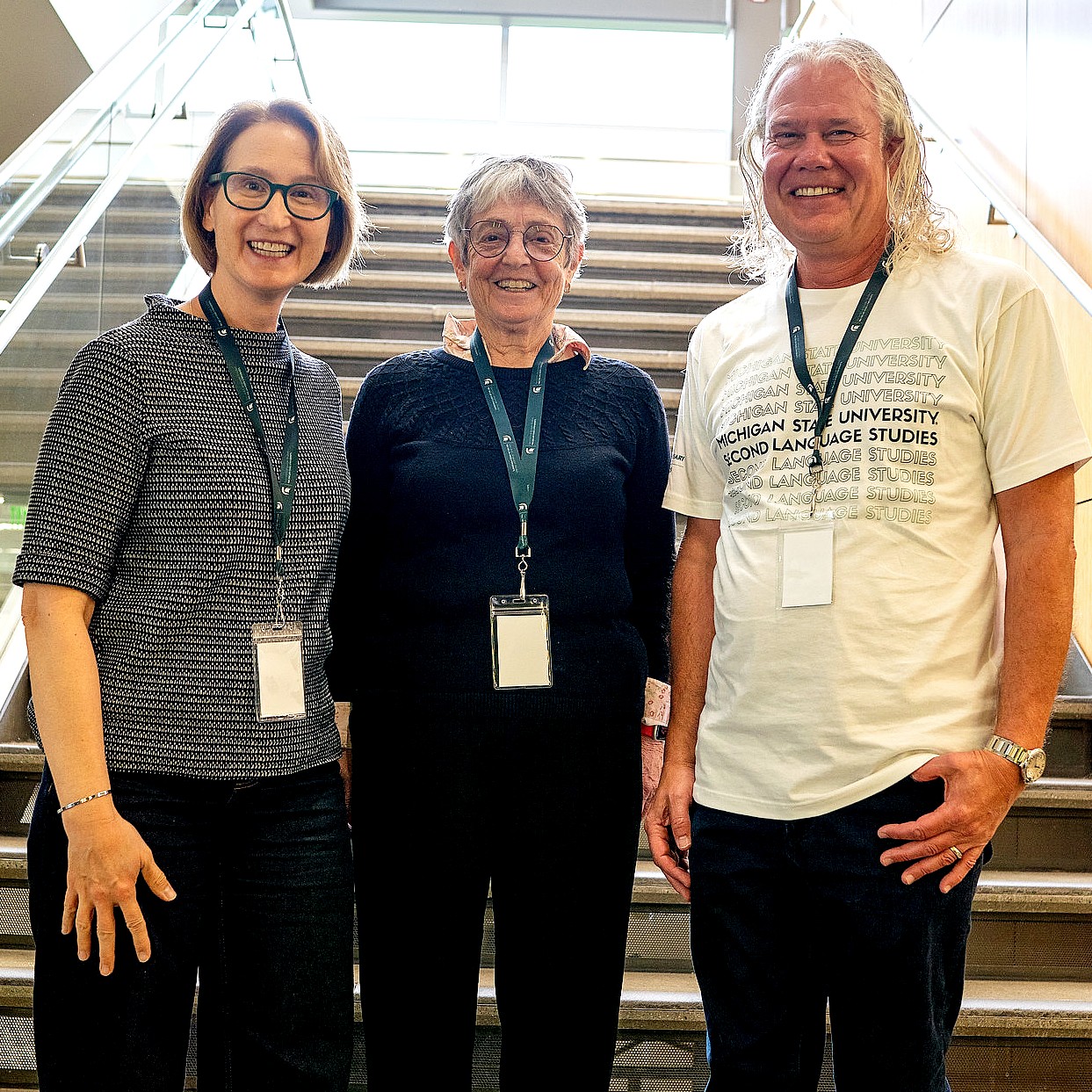 Drs. Paula Winke, Susan Gass, and Shawn Loewen, wearing name badges stand together in front of a staircase at the Second Language Studies 20th anniversary event smiling. 