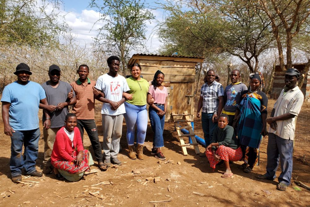 A group of about a dozen people stands in a line outdoors in front of a small wooden coop. The individuals wear a mix of casual clothing, including long skirts, jeans, hats, and patterned fabrics. The ground is dry and dusty, and trees with sparse foliage in the background.