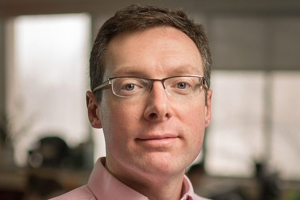 A man with short light brown hair, rectangular glasses, and a light pink collared shirt, indoors with a blurred background