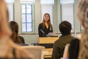 Rachel Zhai stands at a podium holding notes while delivering an oral presentation to an audience seated in a lecture-style room at the Diversity Research Showcase.