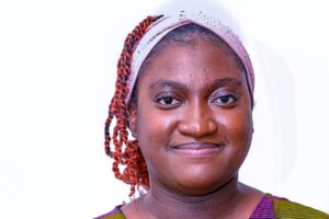 Portrait of a woman facing the camera against a white background, wearing a light headband, with braided hair pulled back and a slight smile.