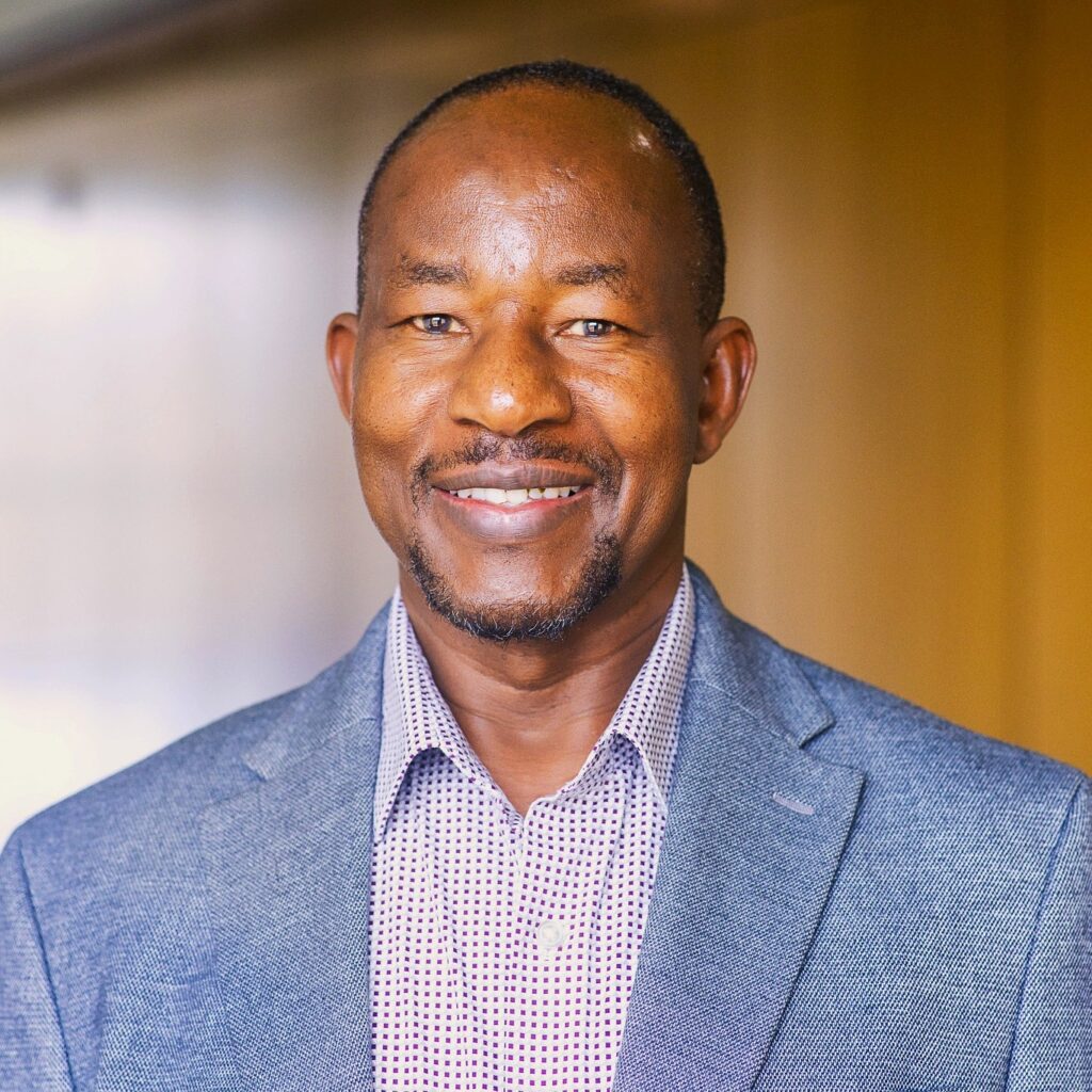 A headshot of Jonathan Choti, a man with short dark hair and a slight goatee, smiling warmly. He is wearing a light blue textured blazer over a white and purple checkered button-down shirt. The background is a blurred, brightly lit hallway with warm yellow and tan tones.