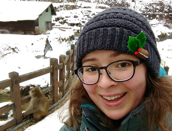 girl with winter hat and glasses standing in front of a snowy background and fence with a monkey on it