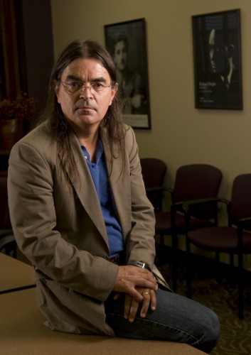 Man with long brown hair and glasses is wearing a brown blazer and blue shirt. He is sitting on a wooden desk with his hands folded over his lap