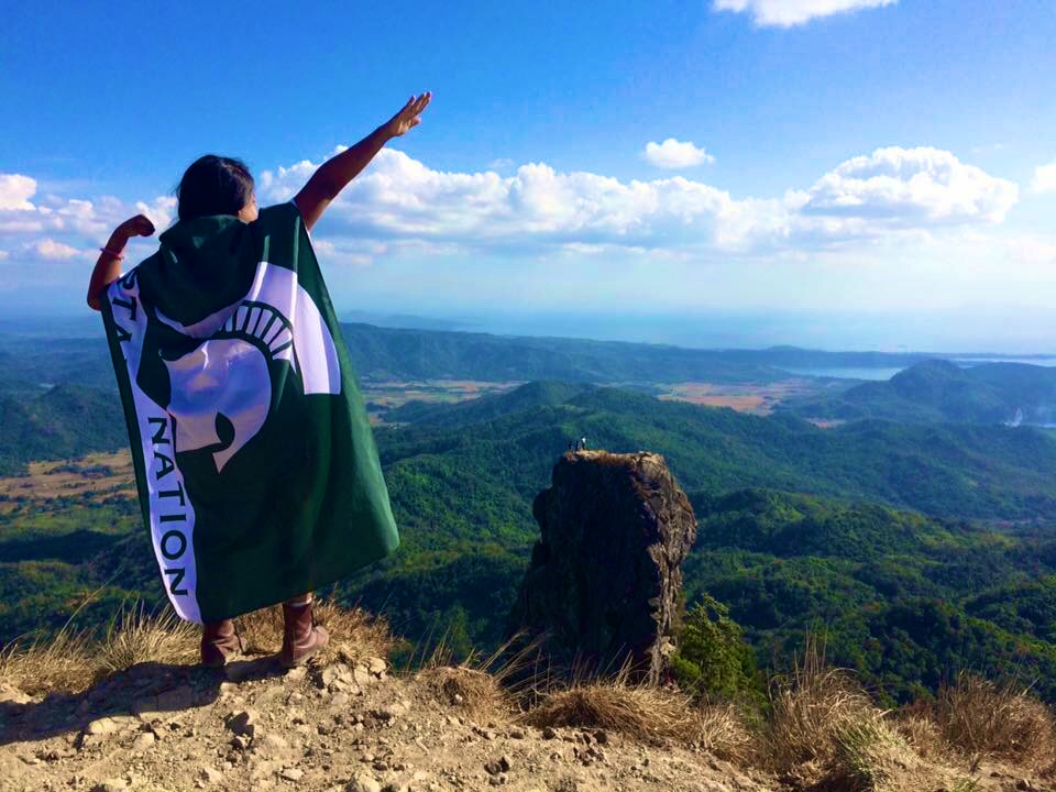 woman with a Michigan State University flag on her back standing on a mountain in front of a clear blue sky