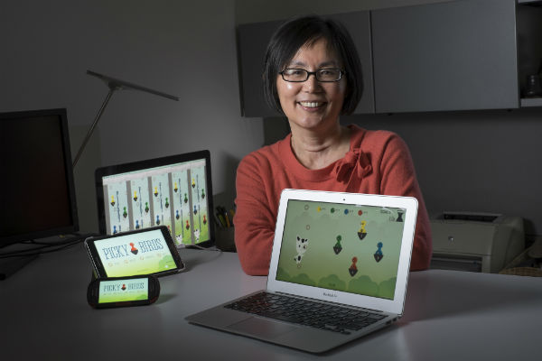 A woman in a red sweater smiling with several electronic devices displaying a game called picky birds
