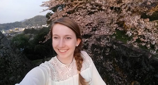 woman wearing a white shirt and taking a selfie in front of scenic mountains and cherry blossom trees in Japan