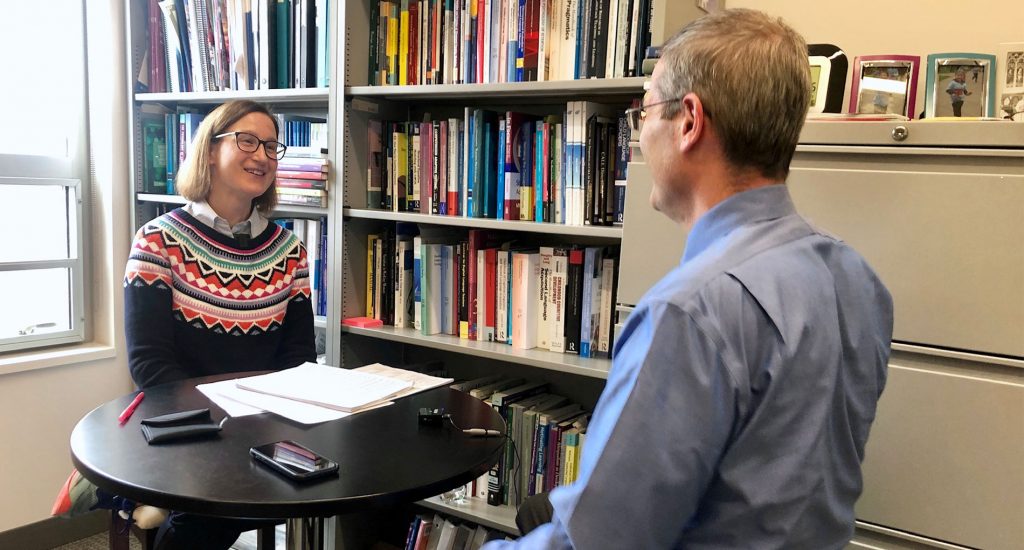 two people talking across the table in front of a book shelf