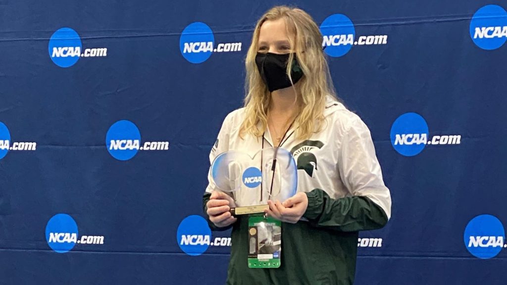Photo of a woman with blonde hair holding an award made of glass shaped in the number "90". She is wearing a black face mask and a white and green windbreaker. Behind her is a blue backdrop with light blue circles and the acronym, NCAA.com 