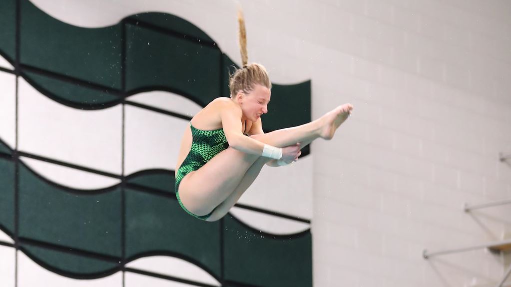 Photo of a woman with blonde hair in a ponytail diving. Her arms are wrapped around her outstretched legs and she is wearing a green one-piece swimsuit