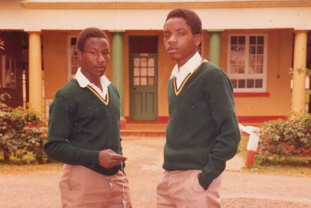 A medium shot of two African American teenagers in matching school uniforms stand in front of a house.