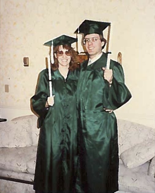 A man and a woman smile in green graduation caps and gowns. They are holding diplomas. 
