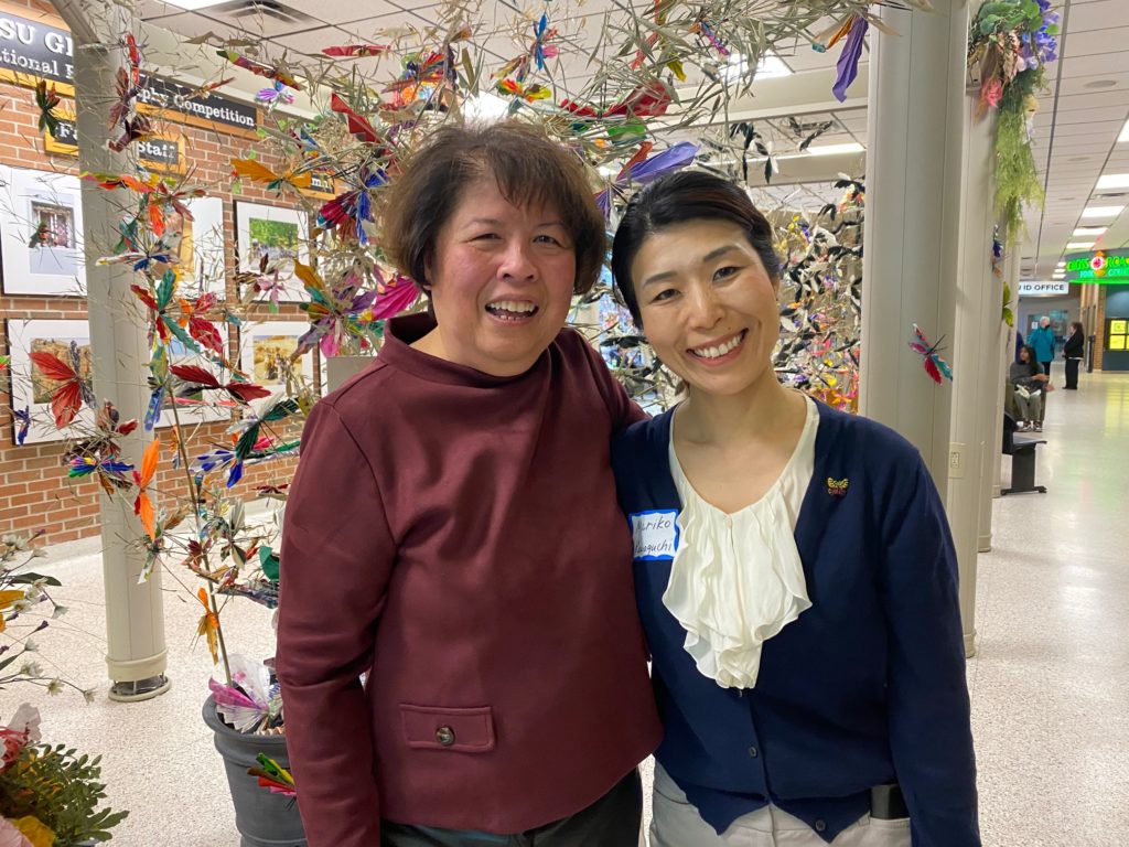 A picture of two women side by side. The woman on the left has short brown hair and wears a red shirt; the woman on the left has black hair pulled back and wears a white and blue shirt. The are posing before an art exhibit. 
