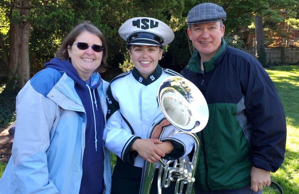 A smiling young woman dressed in green and white marching band garb stands outdoors with a smiling woman to her left and a smiling man to her right.