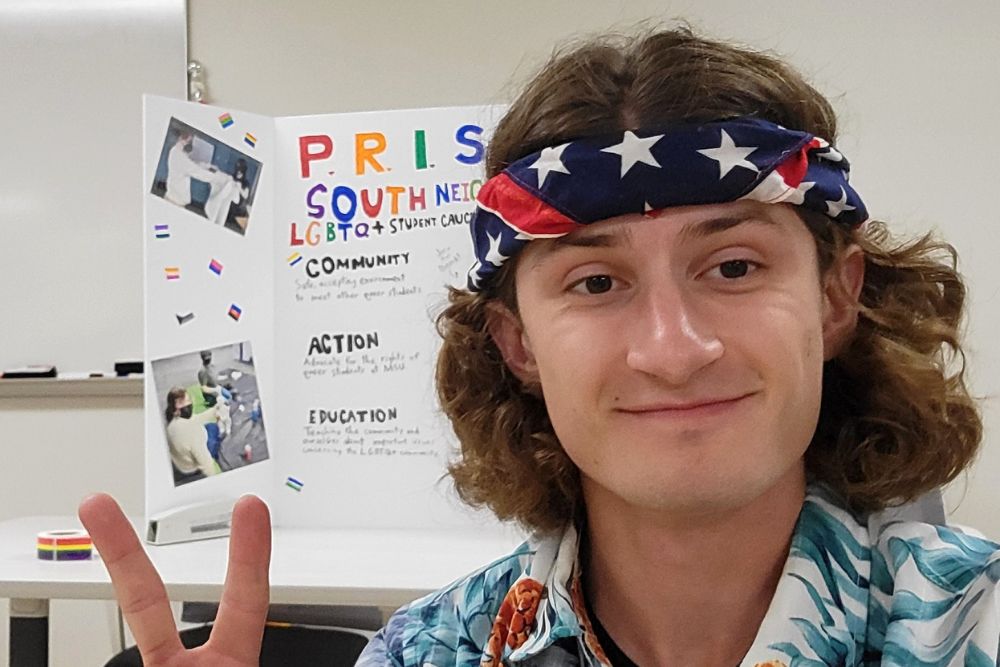 A smiling young man with shoulder-length brown hair and brown eyes wearing an American flag bandanna headband takes a selfie in front of a display board with photos and writing on it.