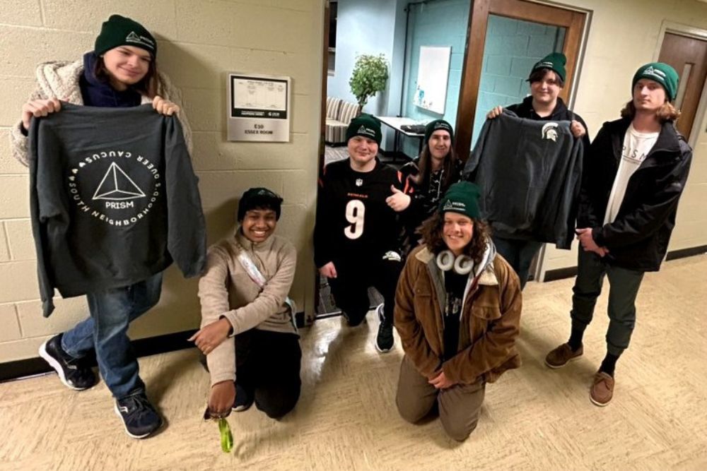 Seven young people sit and stand in a university hallway, all with green hats on. Two people, one at far left and another second to right, hold up different sweatshirts.