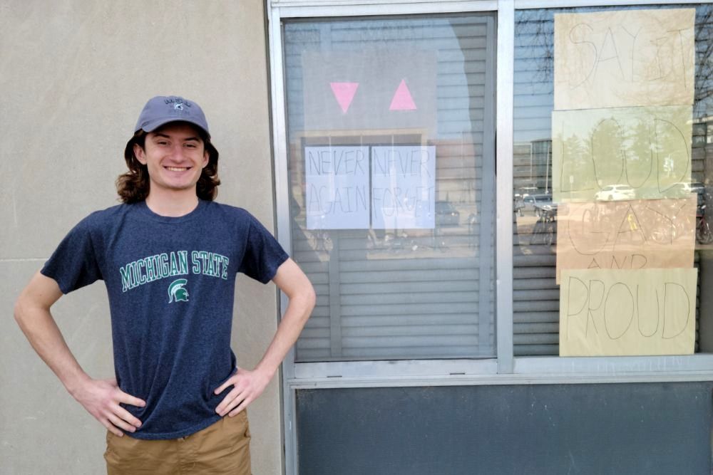 A smiling young man with shoulder-length brown hair and brown eyes wearing a cap stands with his hands on his waist in front of a window that has papers with "never again, never forget" and "say it loud, gay and proud" in all caps taped onto it from inside the building.