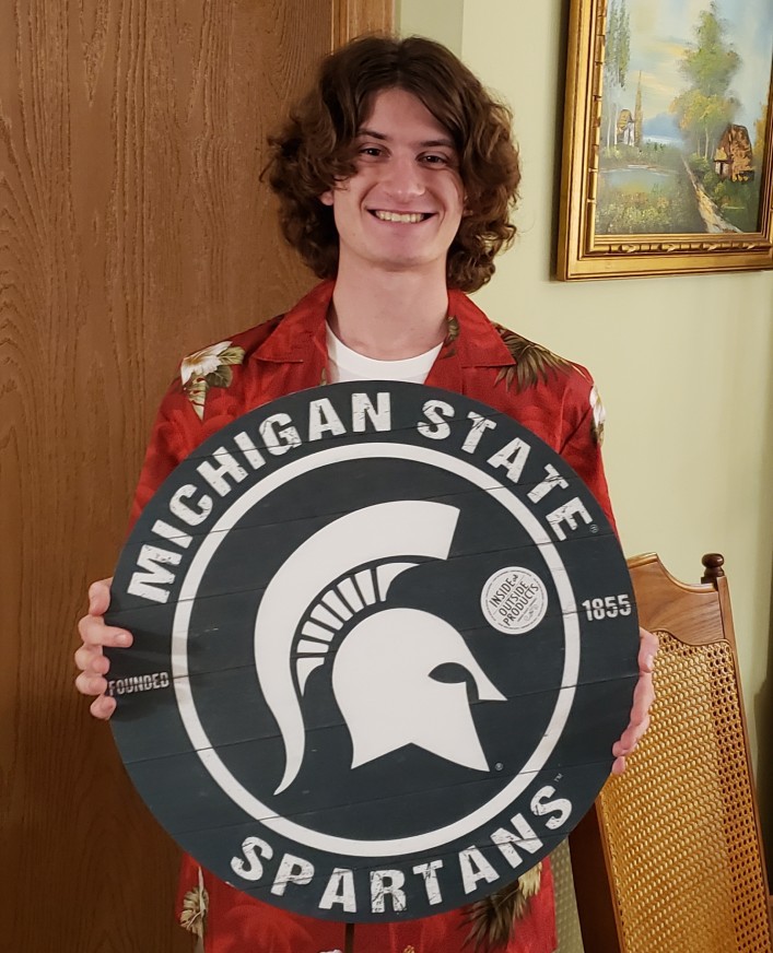 A smiling young man with shoulder-length brown hair and brown eyes wearing a red jacket holds a circle decor piece with "Michigan State Spartans" written on the rim with a large Spartan logo in the middle.