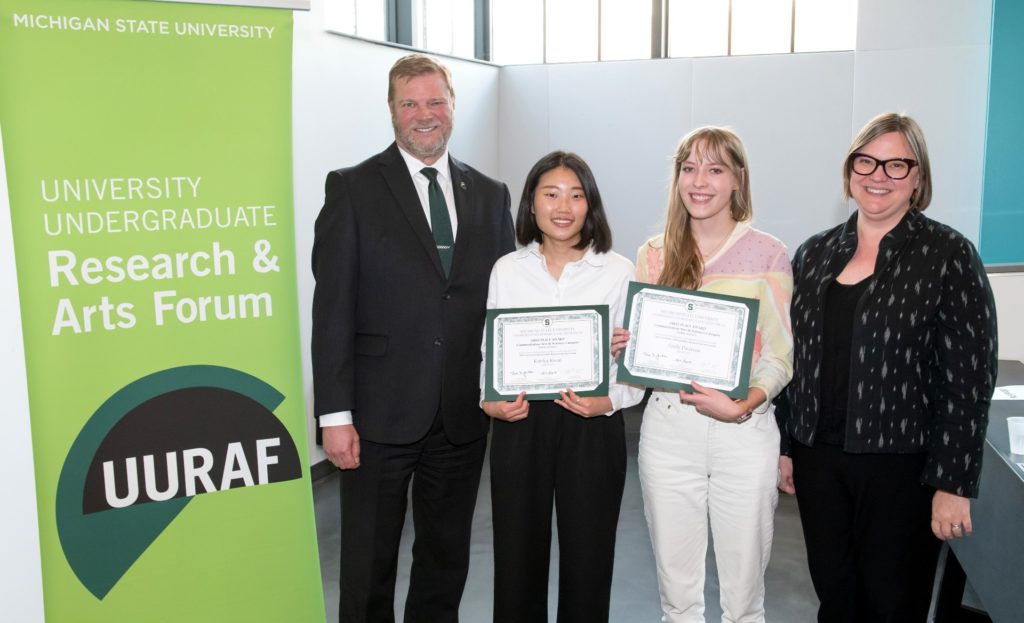Photo with four people, two of who are holding certificates. they are standing next to a banner that says "University Undergraduate Research & Arts Forum." 