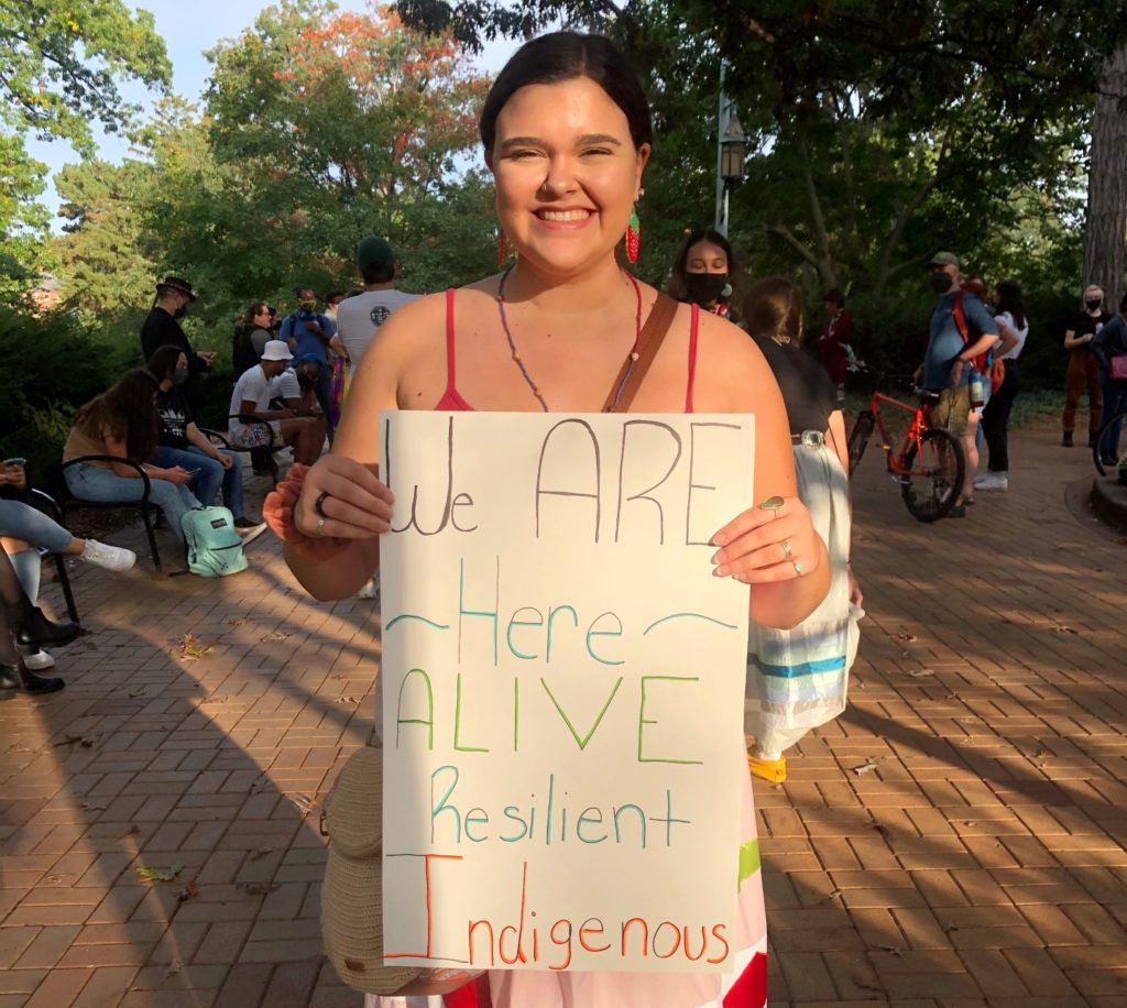 A picture of a woman with black hair wearing a red tank top, holding a sign that reads "We ARE Here; Alive; Resilient; Indigenous"