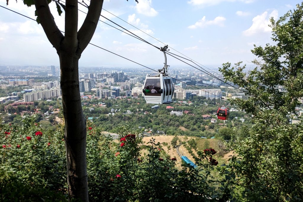 A picture of two cable cars and trees on a sunny day. Beyond the cable cars, in the background, there are rolling hills, houses, and a city. 